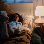 Dziewczynka odpoczywa w łóżku z misiem przy lampce nocnej